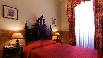Una habitación de hotel con una cama grande y ornamentada, dos mesitas de noche con lámparas, un cuadro enmarcado en la pared y una ventana con cortinas.