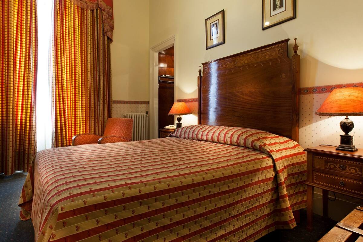 A hotel room with a large bed, a wooden headboard, two bedside tables with lamps, a chair, and a radiator.