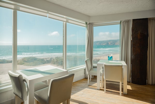 A room with a large window overlooking the ocean, a table with chairs, and a wooden floor.