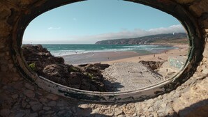 Une vue sur la plage, encadrée par une arche de pierre, avec un premier plan rocheux et un ciel dégagé.
