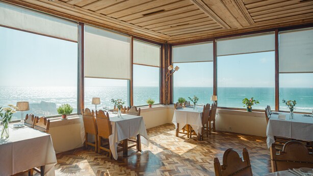 Un espace repas doté d’un plancher en bois et de grandes fenêtres donnant sur la mer. Les tables sont dressées avec des nappes blanches et des chaises.