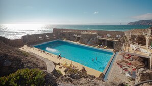 Un espace piscine offrant une vue sur la mer, entouré de murs en pierre et agrémenté de chaises longues et de parasols.