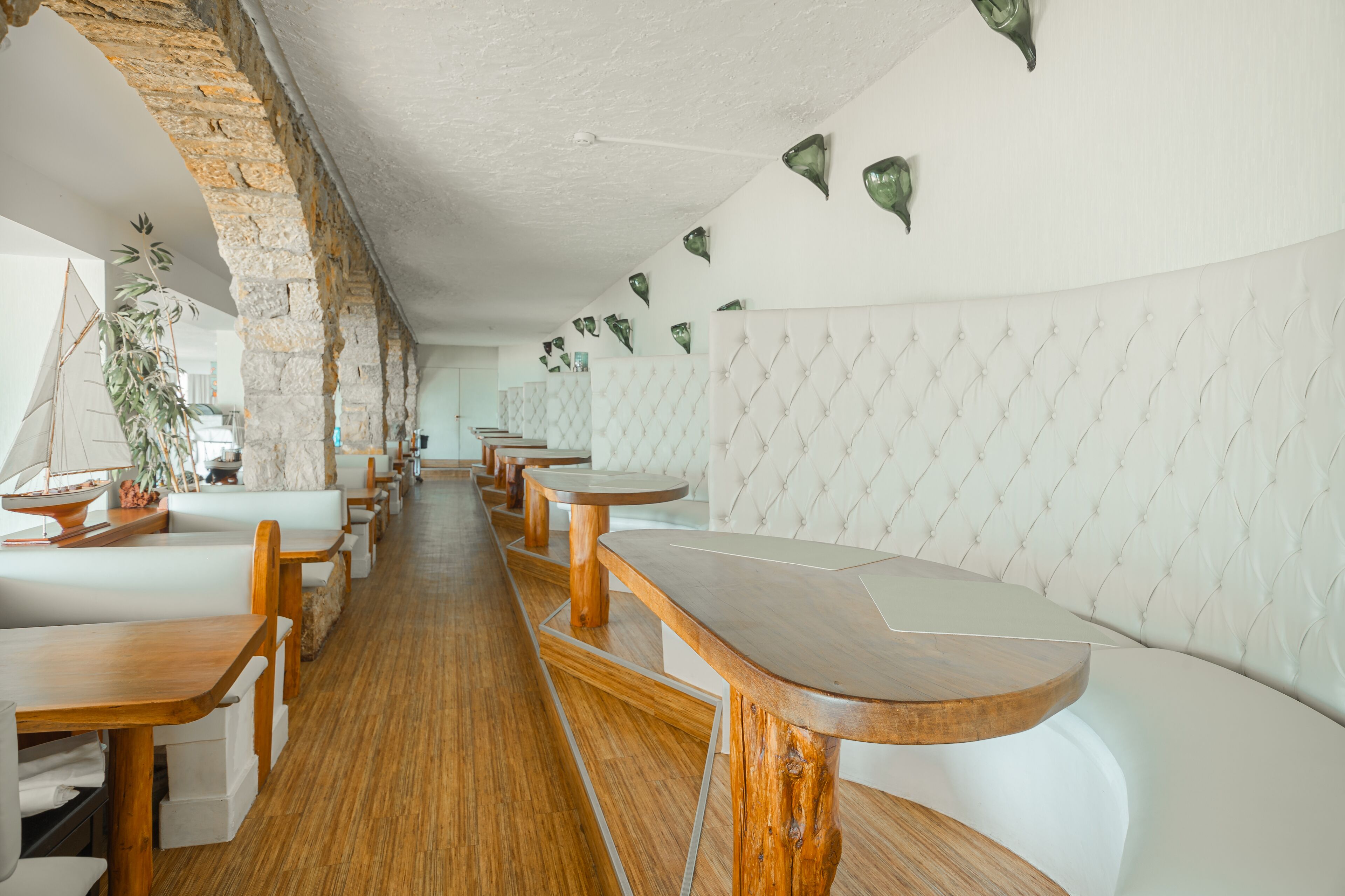 Un restaurante con mesas de madera, asientos blancos acolchados, paredes de ladrillo y hojas verdes colgantes.