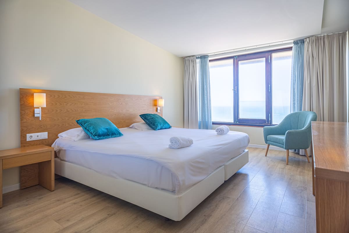 Une chambre d’hôtel avec un grand lit, des tables de chevet, une chaise et une vue sur la mer par la fenêtre.
