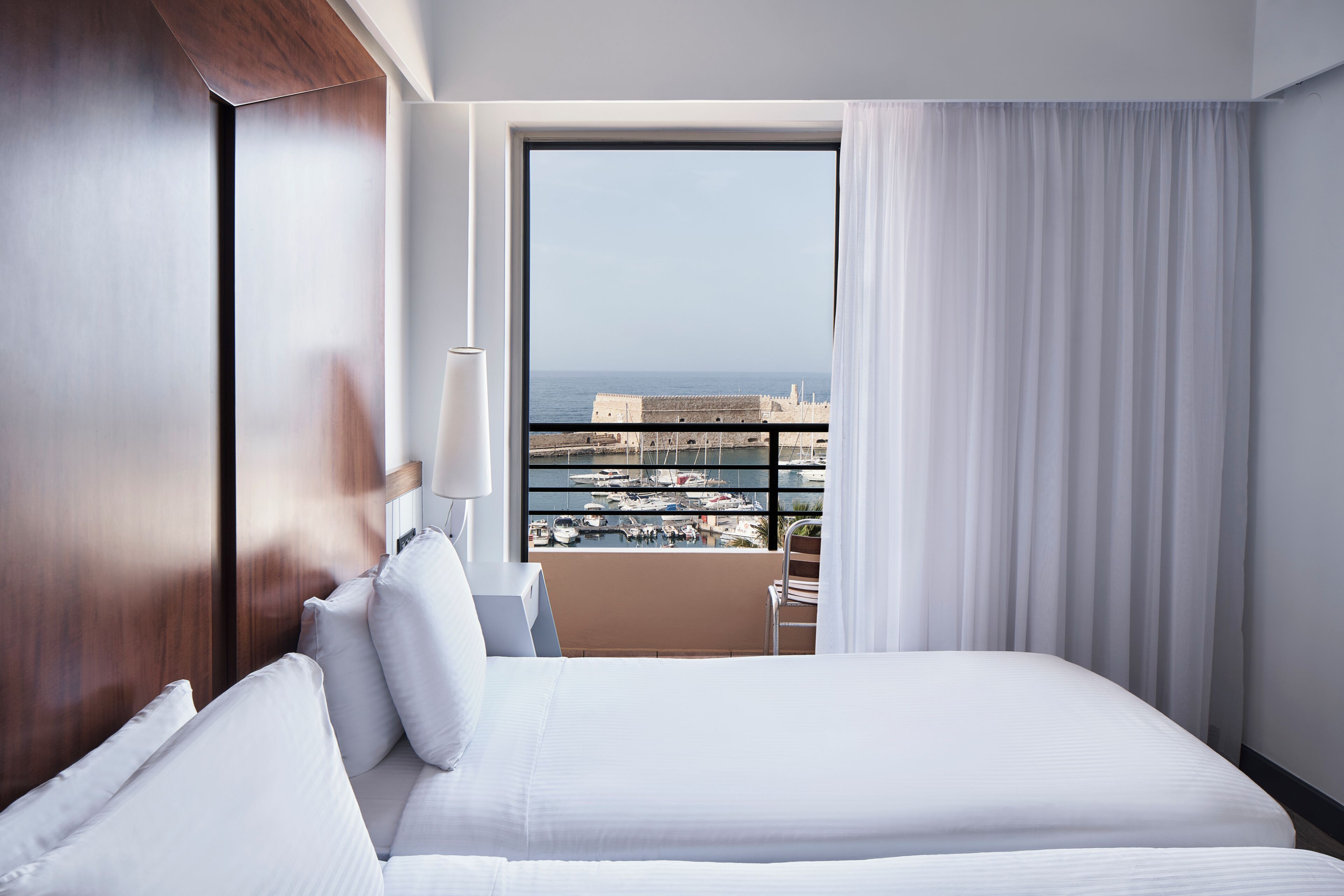 Habitación de hotel con una cama grande, una silla, un escritorio y vistas al mar y a las embarcaciones.
