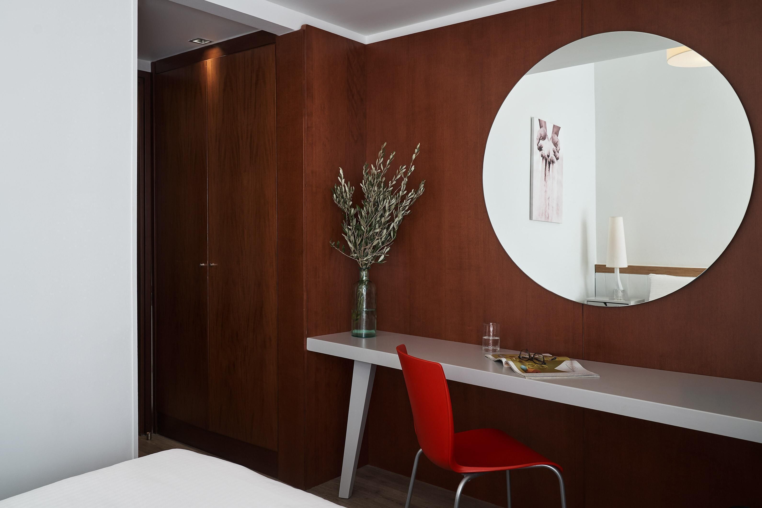 A modern room with a wooden wardrobe, a white desk with a red chair, a round mirror, and a vase with dried plants.
