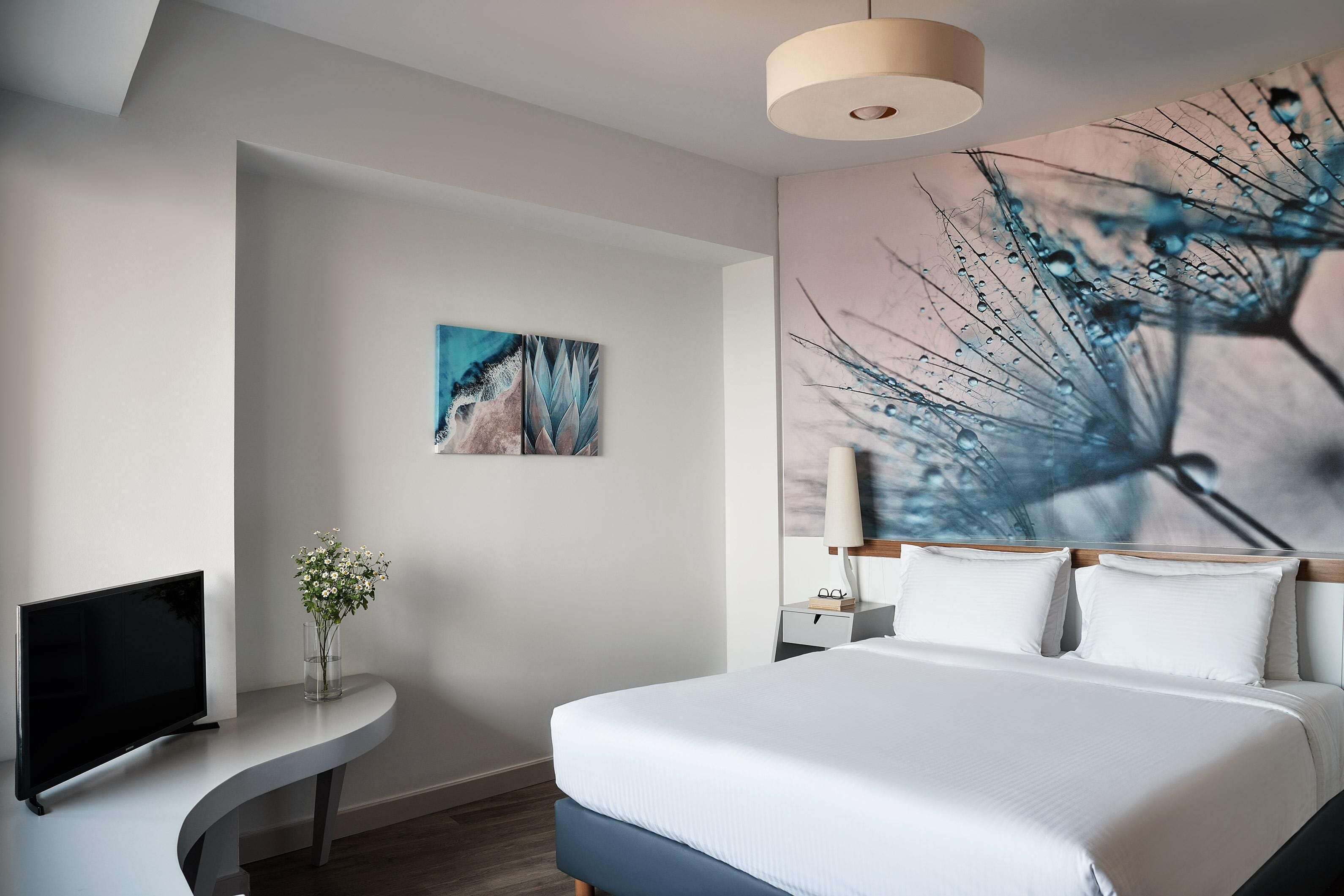 Una moderna camera d'albergo con un letto grande, una televisione a schermo piatto, un comodino con un vaso di fiori e un grande quadro astratto appeso al muro.