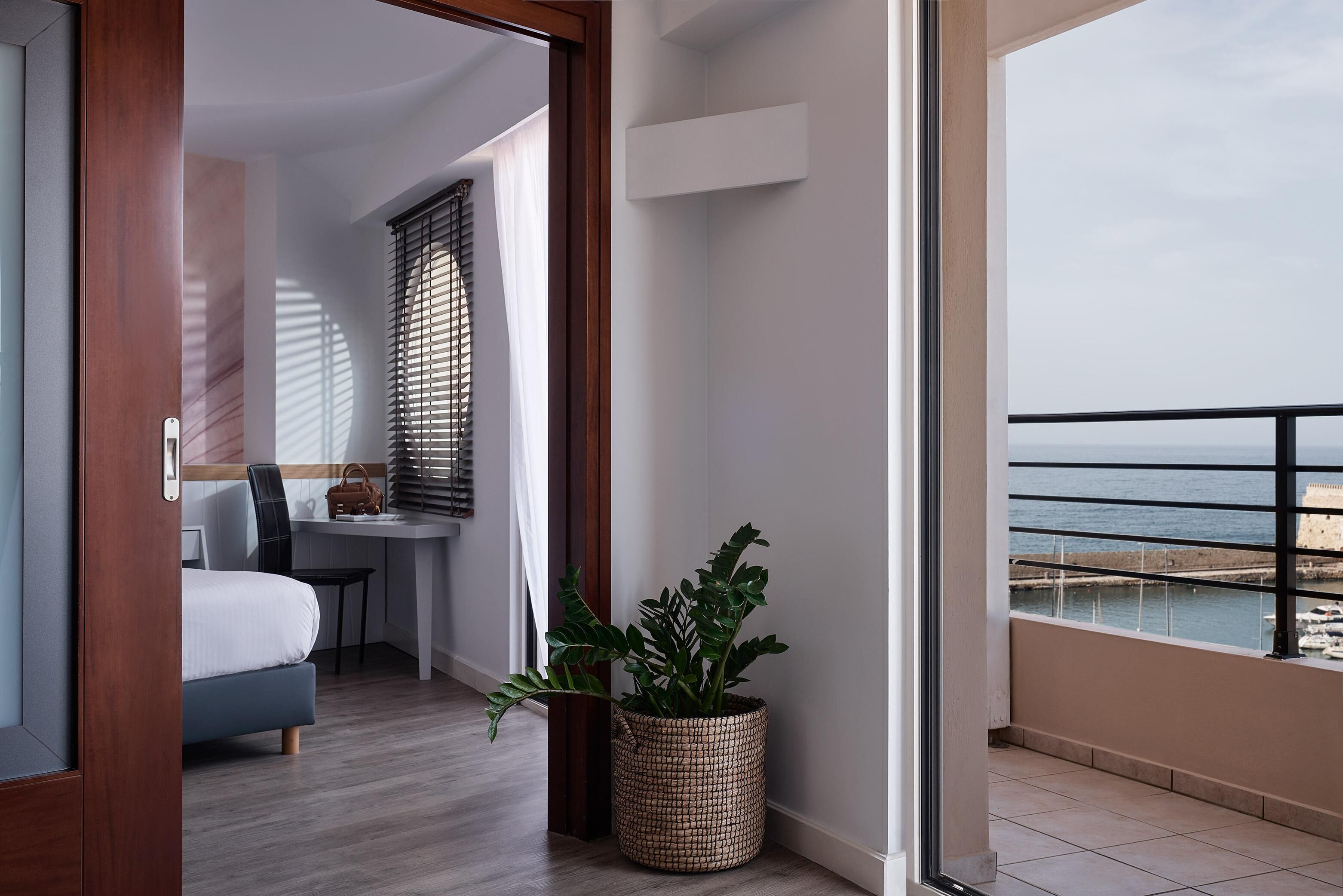 A modern hotel room with a balcony, a bed, a desk, and a potted plant.