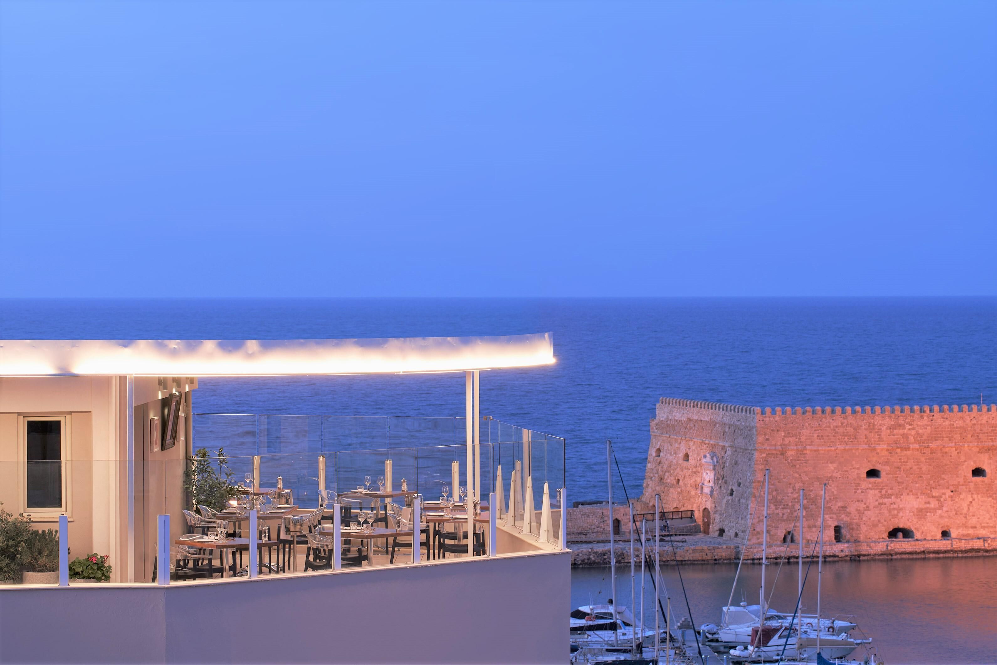 Un ristorante sul mare con posti a sedere all'aperto, affaccio sul mare, un’antica muraglia in pietra e barche sullo sfondo.