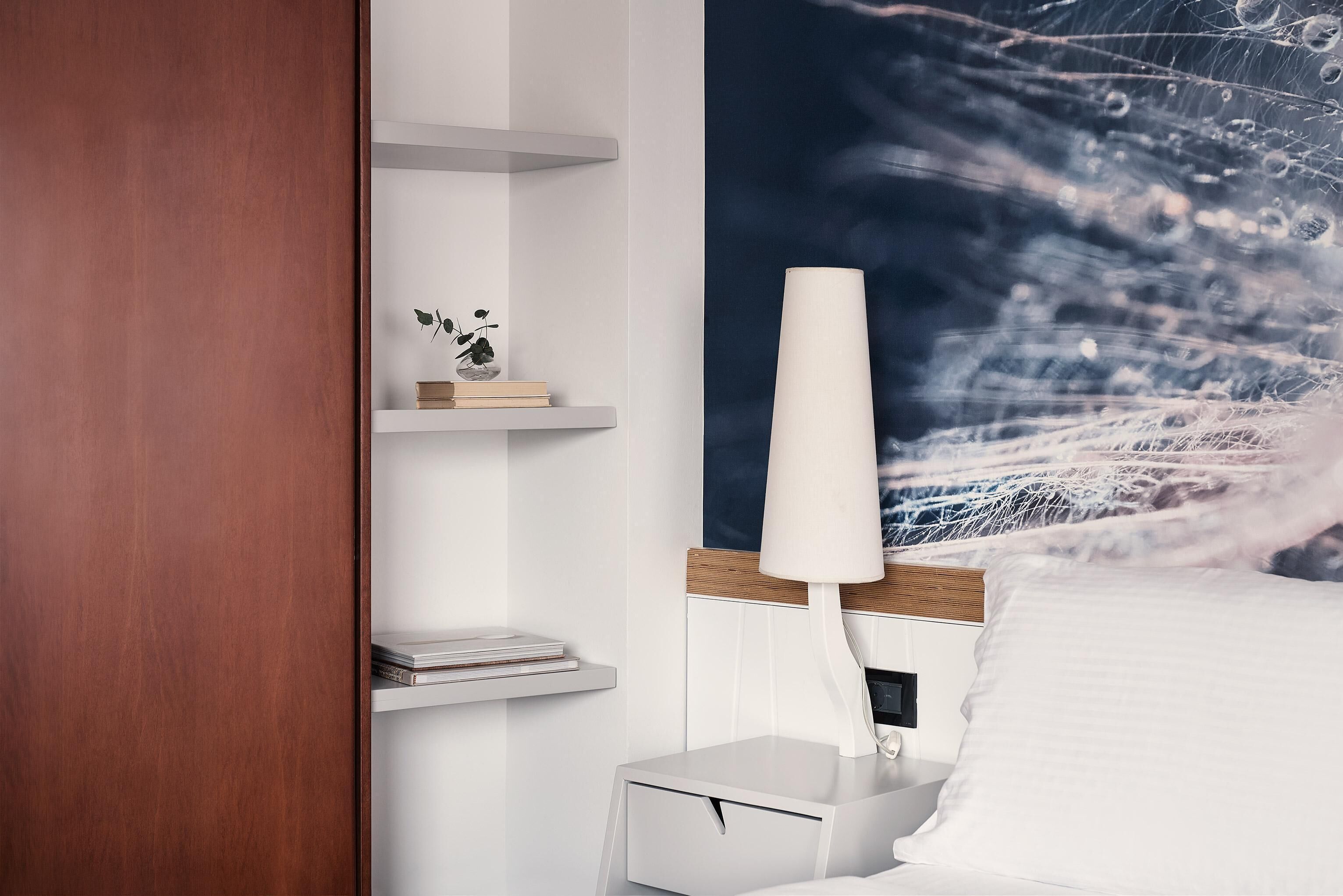 Une chambre avec une table de chevet blanche, une table de nuit avec une lampe, un lit avec une literie blanche et un mur orné d’un tableau abstrait bleu.