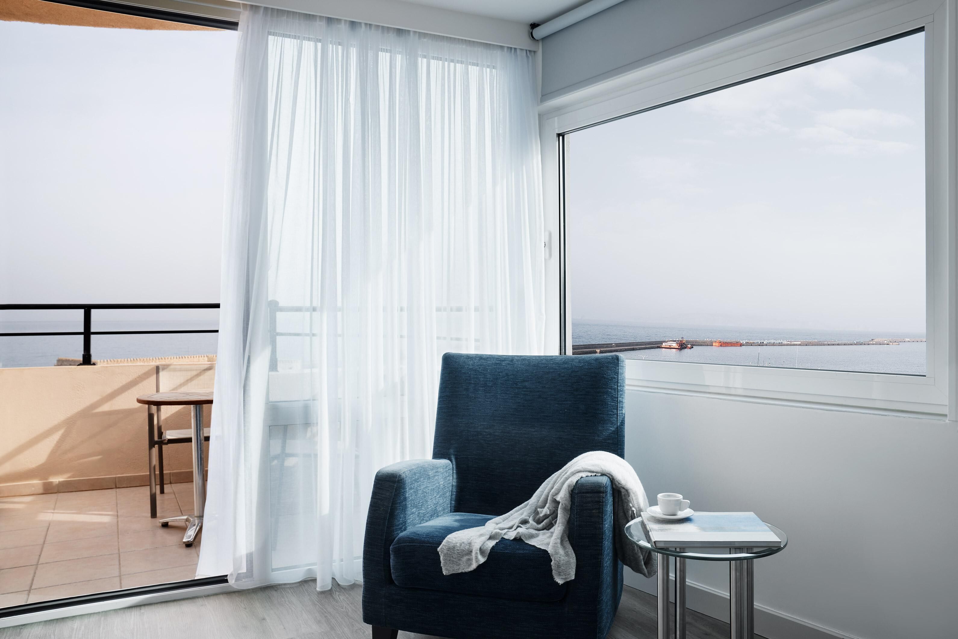 Ein Balkon mit Stuhl und Tisch, Blick auf das Meer und ein großes Fenster mit weißen Vorhängen.