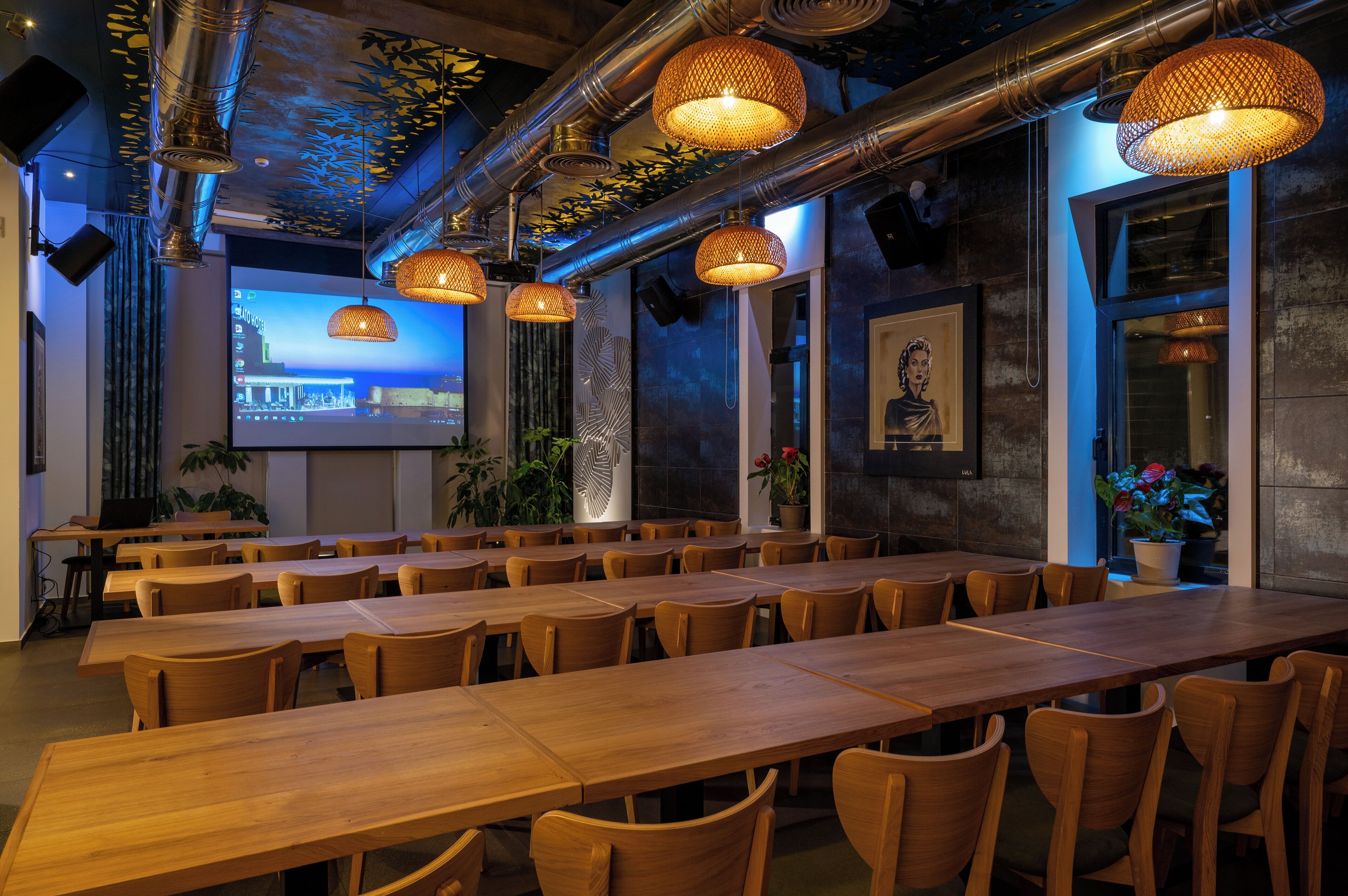 A restaurant with long wooden tables and chairs, hanging wicker lamps, and a projection screen on the wall.