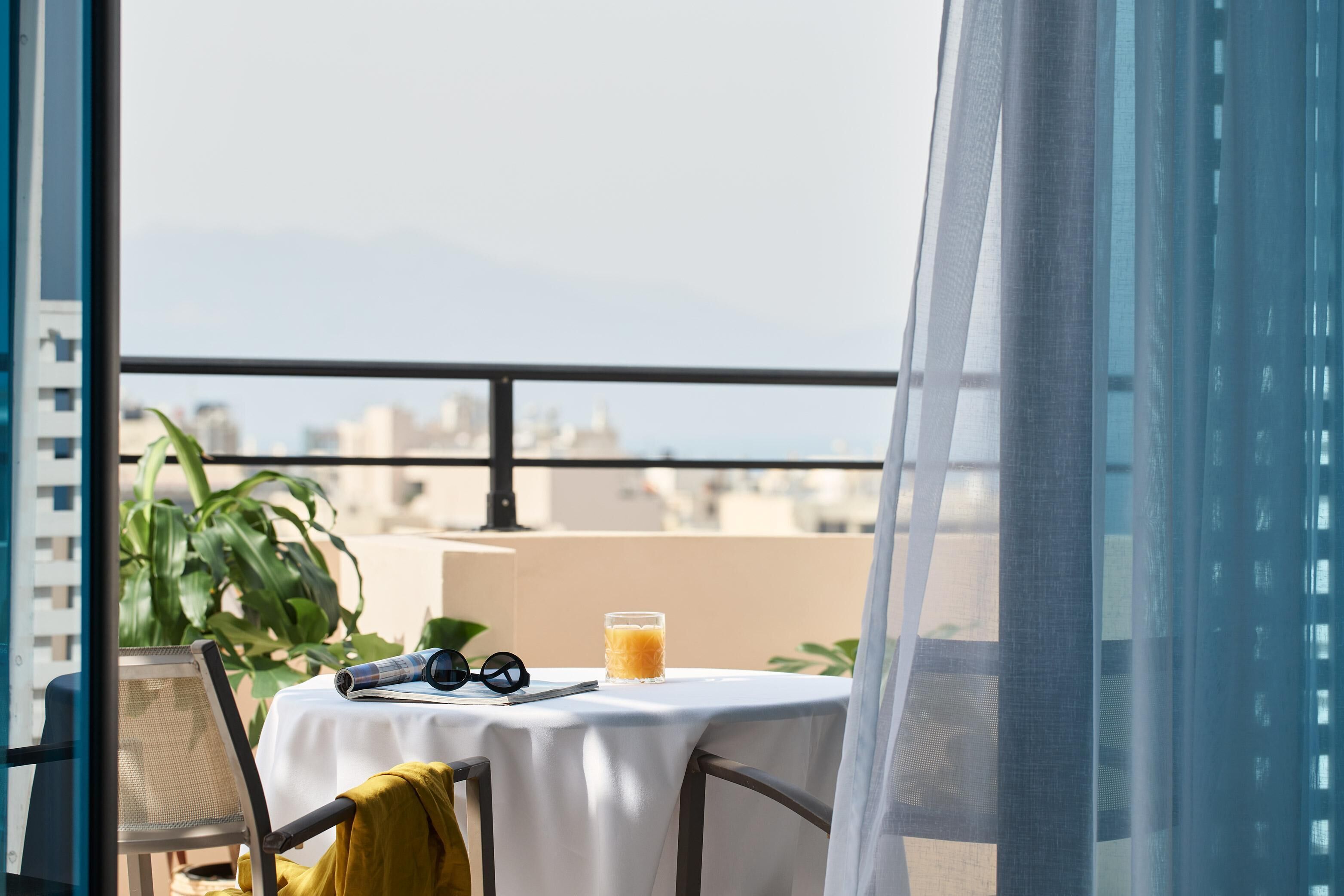 Ein Tisch mit weißem Tuch, ein Glas Orangensaft und eine Sonnenbrille auf einem Balkon mit Blick auf die Stadt.