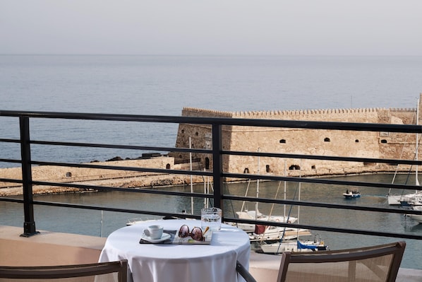 Une table avec un verre d’eau, une tasse et des lunettes de soleil, sur un balcon donnant sur un port où se trouvent des bateaux et un fort historique.