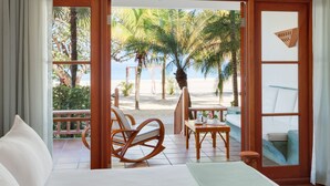 A hotel room with a bed, a wooden chair, a small table, and a view of the beach and palm trees.