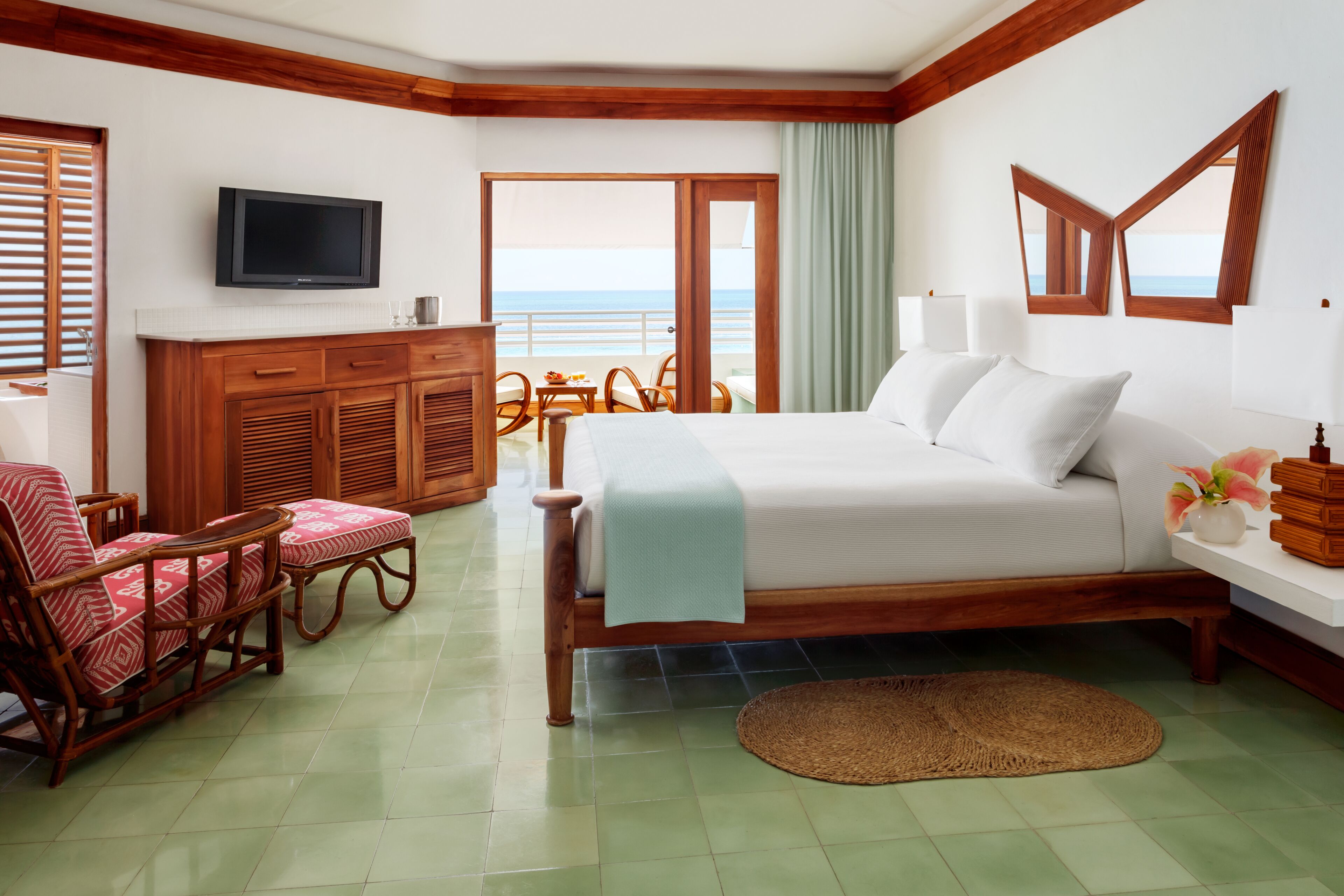 A hotel room with a bed, a television, a wooden dresser, wicker chairs, and a view of the ocean.