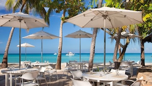 Un restaurante frente al mar con mesas y sillas blancas, sombrillas grandes y vistas al océano.