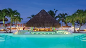 Zona de piscina con bar, palmeras y una estructura de techo de paja.