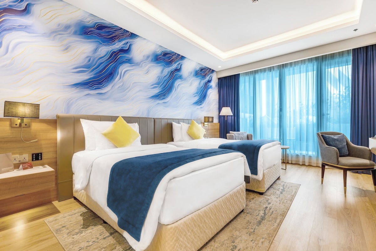 A modern hotel room with two beds, a large abstract mural, a view of the outdoors through sheer curtains, and a wooden floor.