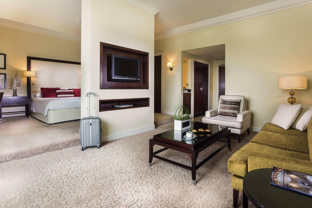 A hotel room with a bed, a TV mounted on the wall, a sofa, a coffee table, and a suitcase.