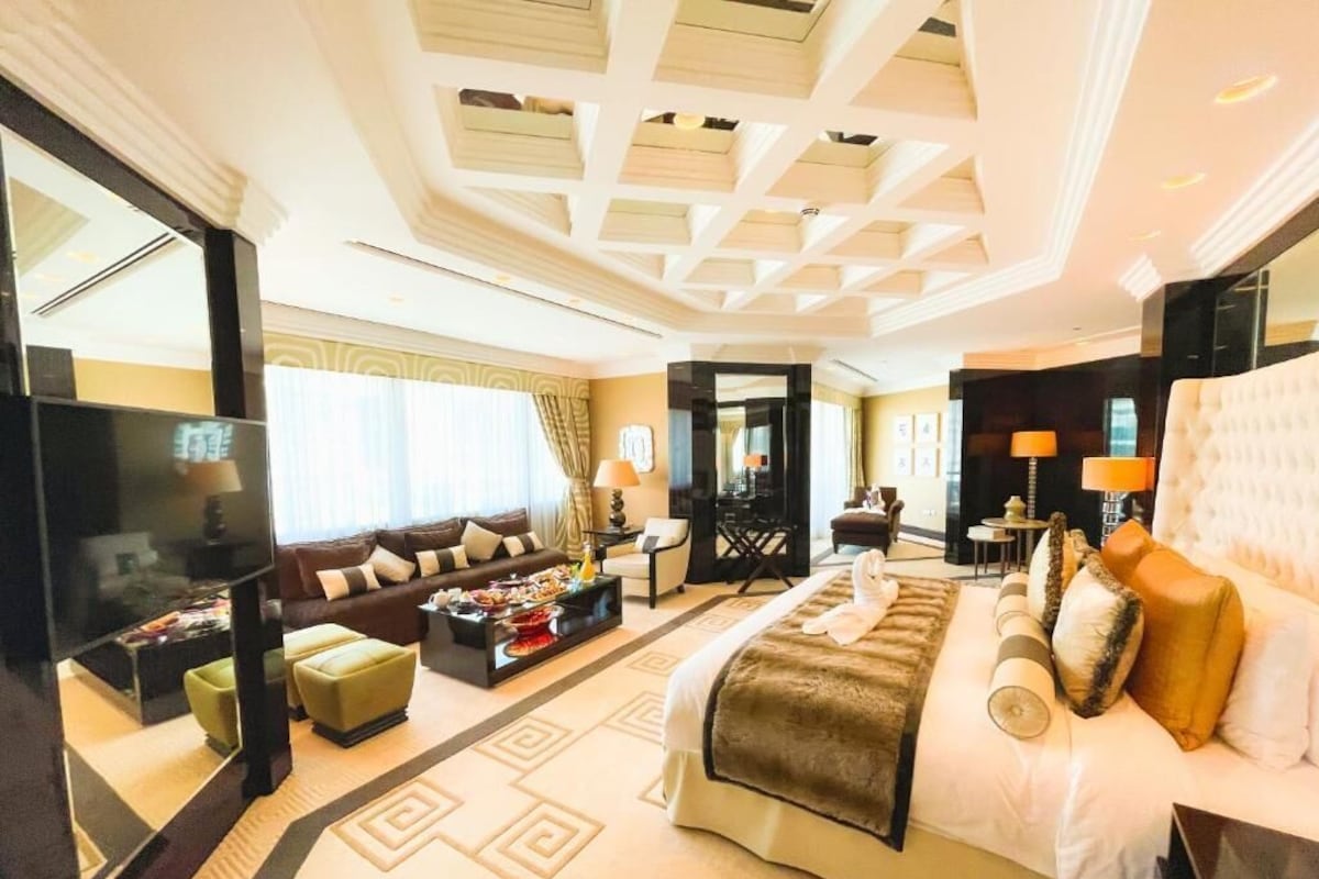 A spacious hotel room with a large bed, a sofa, a dining table, and a TV.