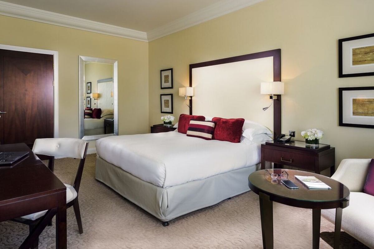 A hotel room with a large bed, a desk, a chair, a round coffee table, and a mirror.