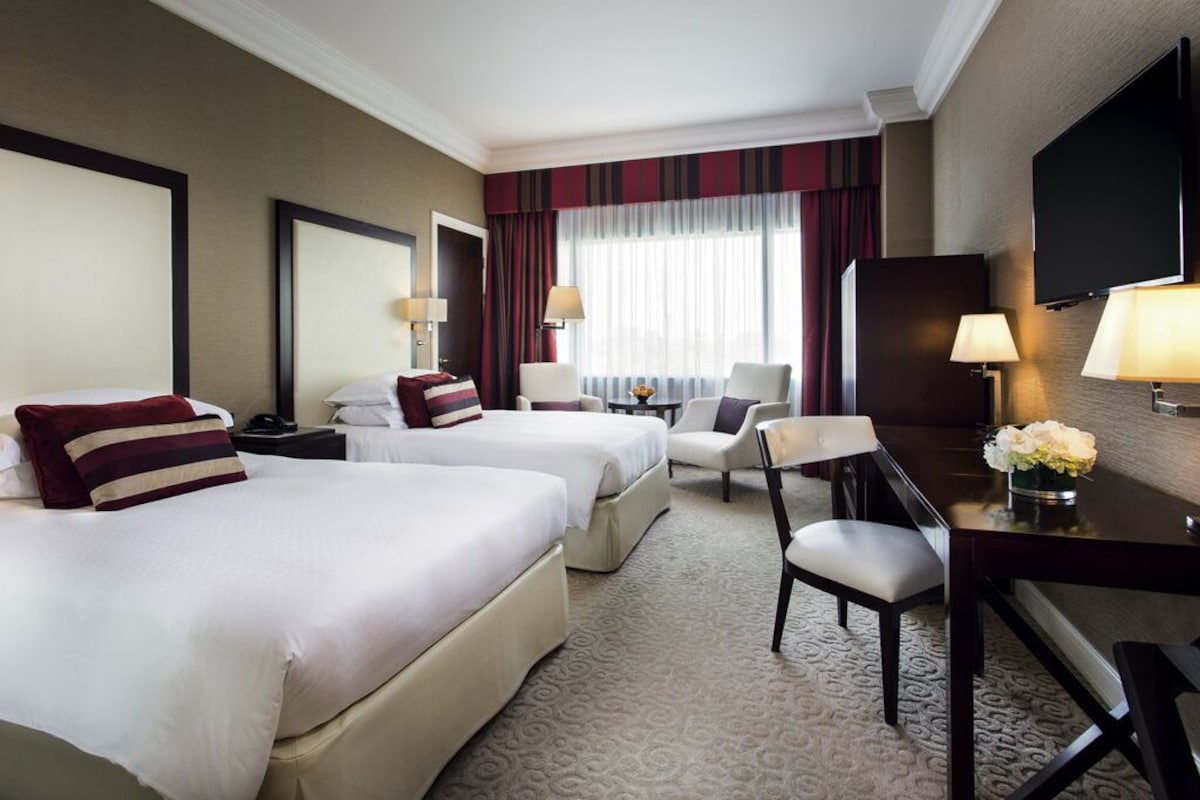 A hotel room with two beds, a desk, a chair, a TV, and a window with curtains.