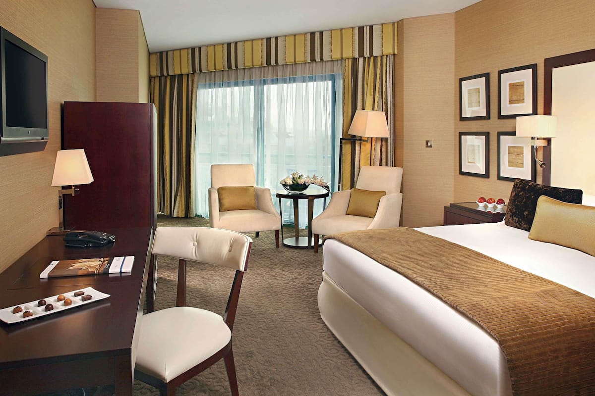 A hotel room with a bed, two armchairs, a desk with a lamp, a television, and a window with curtains.