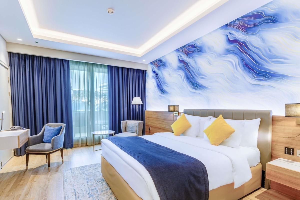 A hotel room with a bed, a desk, a chair, a lamp, and a large abstract mural on the wall.