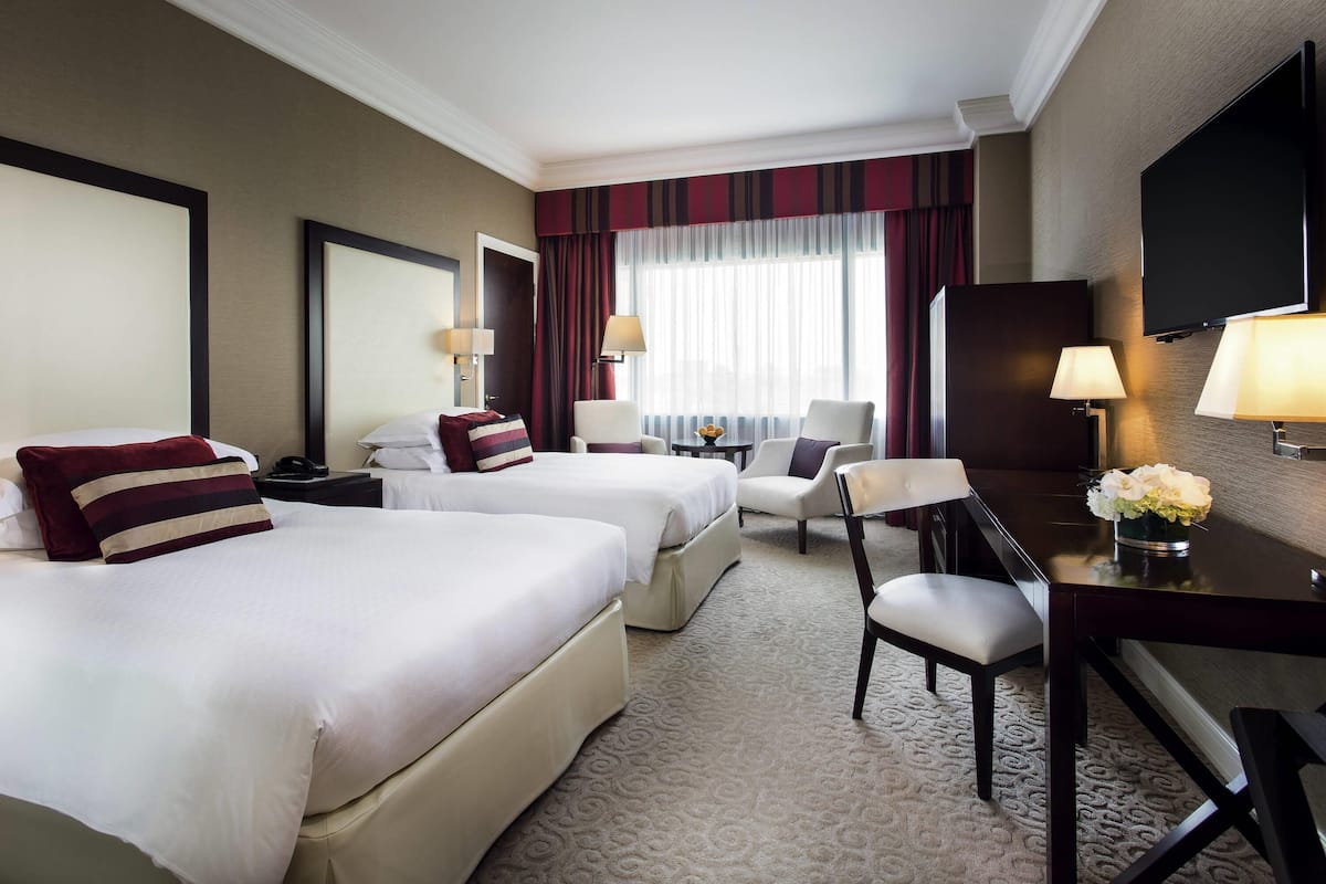 A hotel room with two beds, a desk, a chair, a TV, and a large window with curtains.