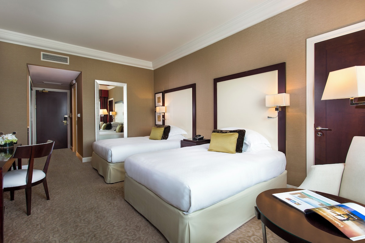 A hotel room with two beds, a desk, a chair, and a table with a magazine.