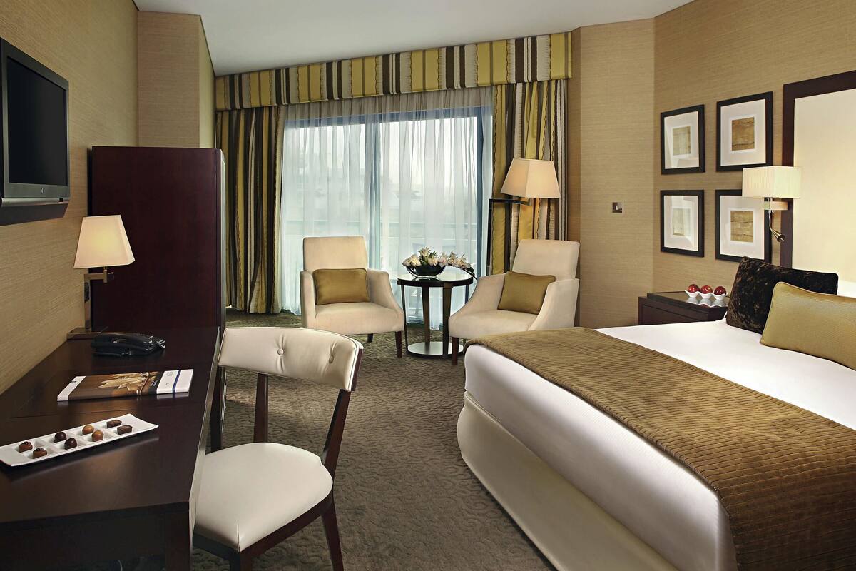 A hotel room with a bed, two armchairs, a desk with a lamp, a television, and a window with curtains.