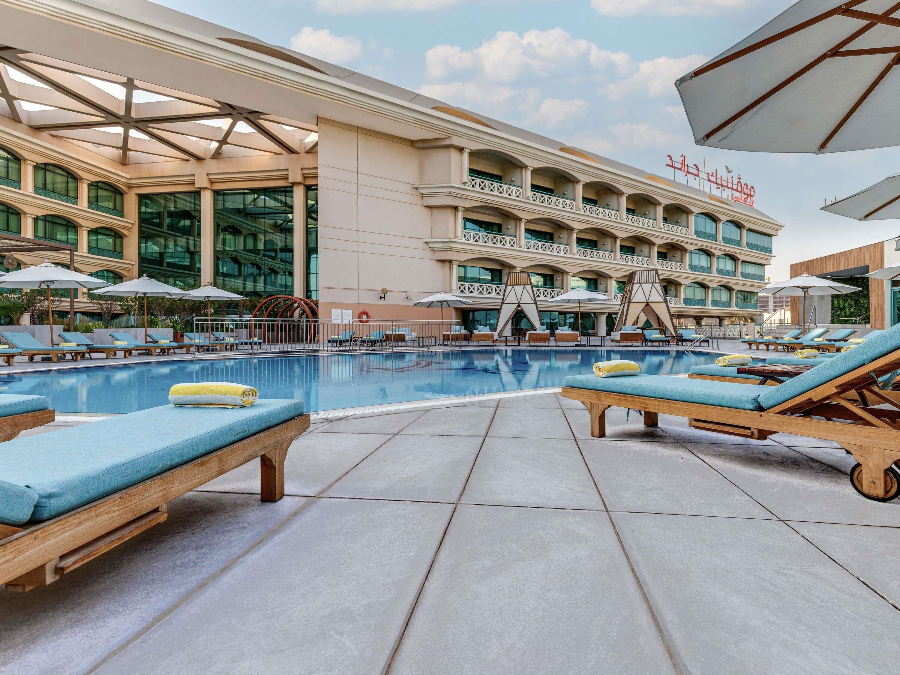 A hotel pool area with sun loungers and umbrellas, a large pool, and a building with balconies and large windows.