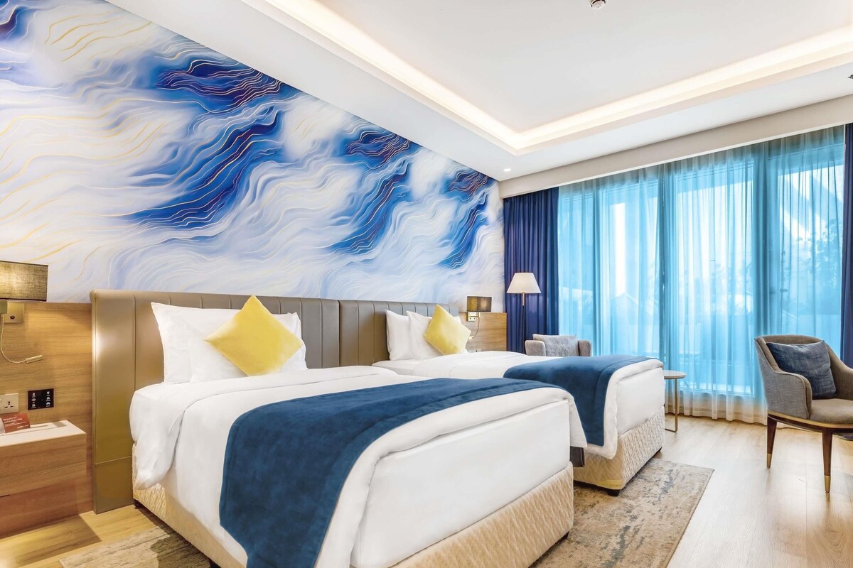 A hotel room with two beds, a large abstract mural, a desk, a chair, and a view of the outdoors through sheer curtains.
