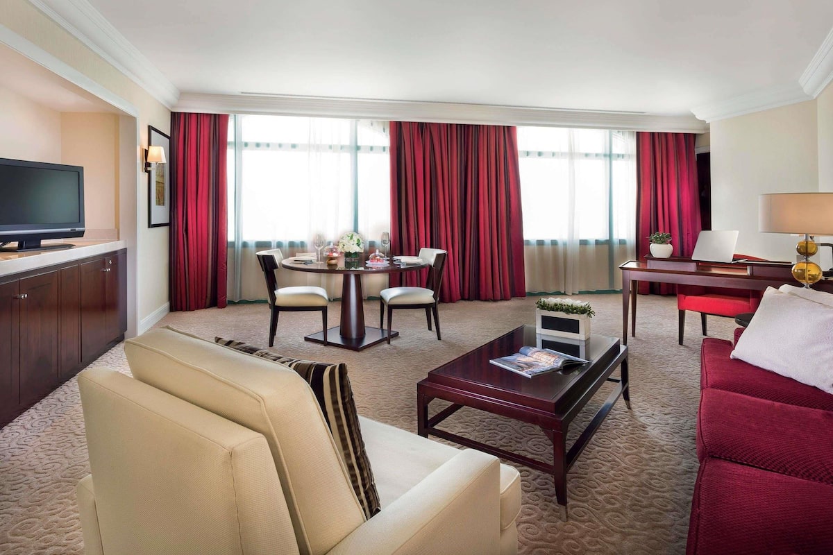 A hotel room with a living area, a dining table with chairs, a sofa, a coffee table, a TV, and a desk with a lamp.