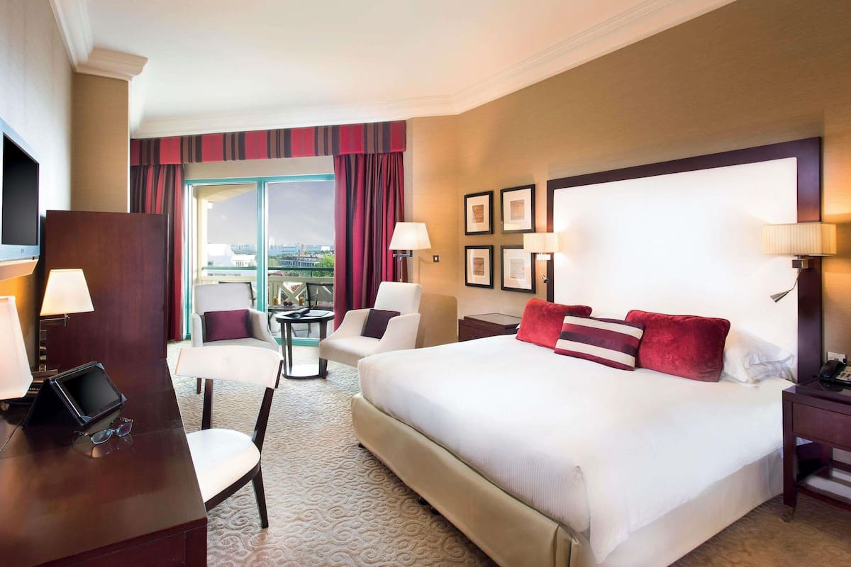 A hotel room with a large bed, two armchairs, a desk with a laptop, and a view of the city through the window.