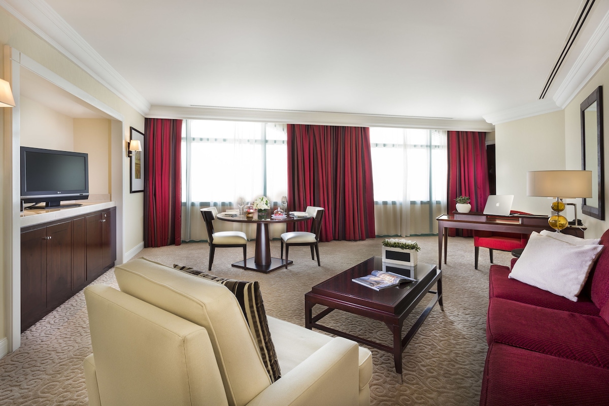 A hotel room with a living area, a dining table, a TV, and a desk with a lamp.