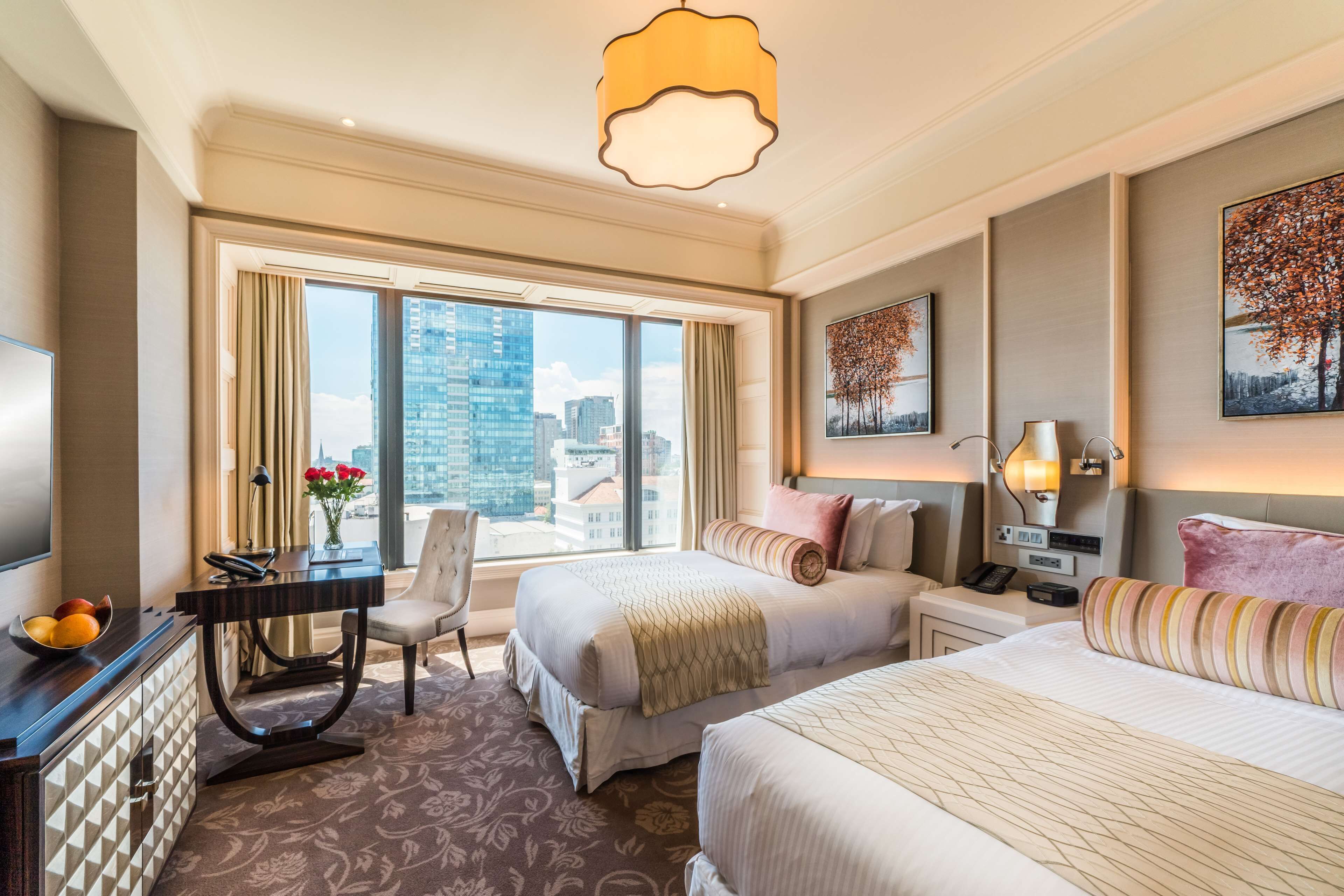 A hotel room with two beds, a desk, a chair, a large window with a city view, and a chandelier.