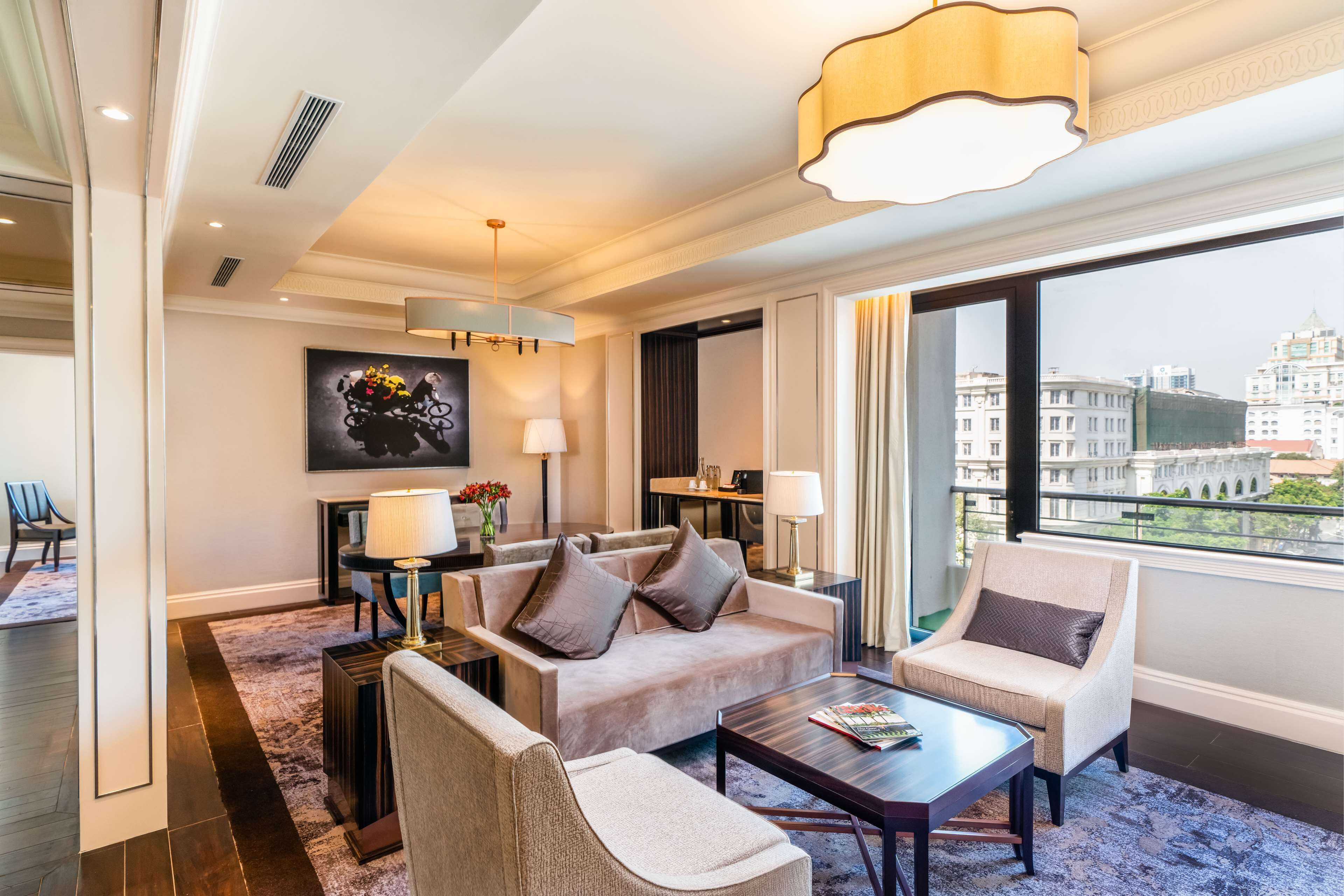 A modern hotel room with a sofa, armchairs, a coffee table, and a large window offering a city view.