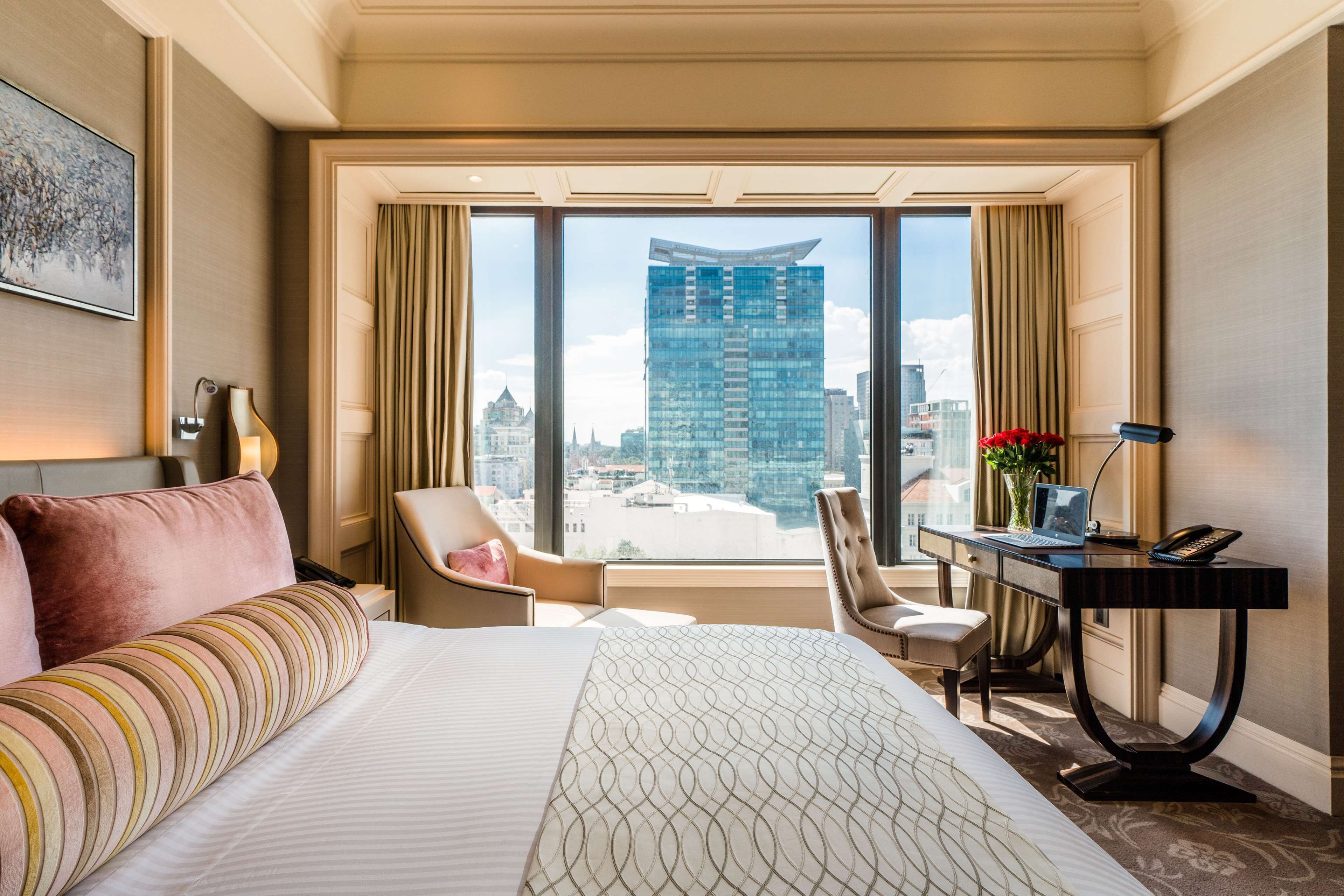 A hotel room with a large bed, a desk with a laptop, a chair, and a view of a cityscape through large windows.