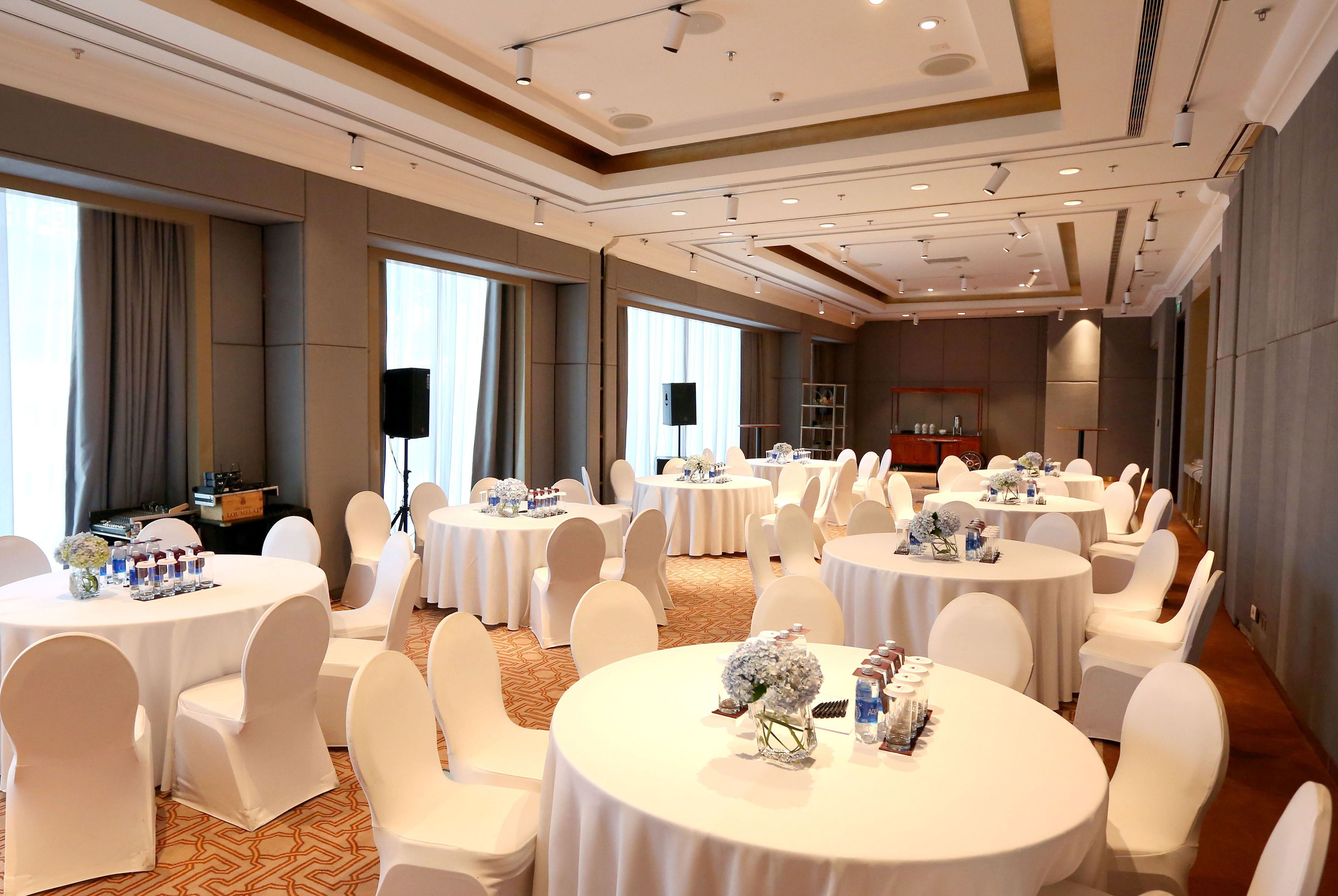 Une salle de conférence d’hôtel avec plusieurs tables rondes, des chaises blanches et une estrade.