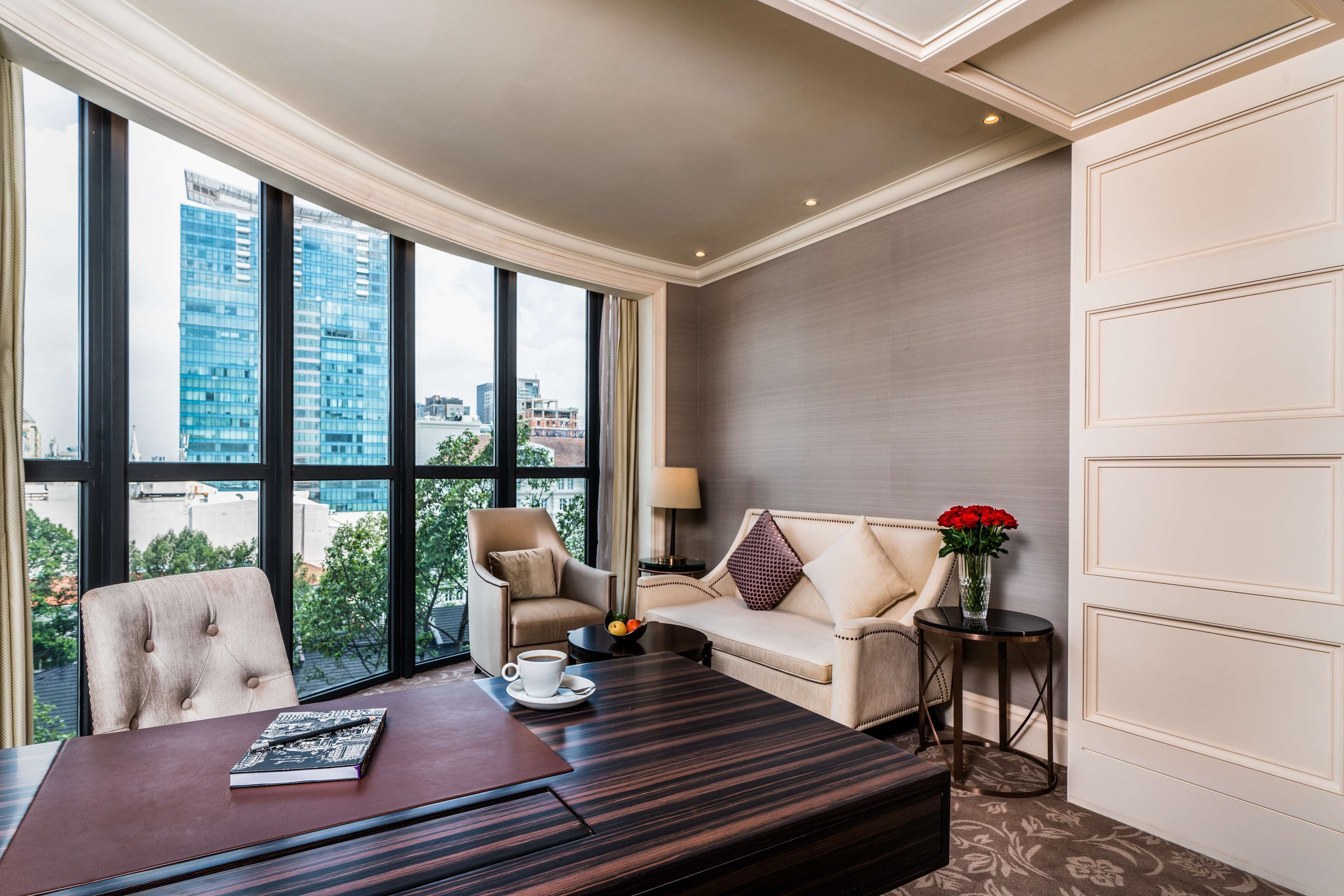 A modern hotel room with a large window, a sofa, a chair, a coffee table, and a small side table with a vase of flowers.