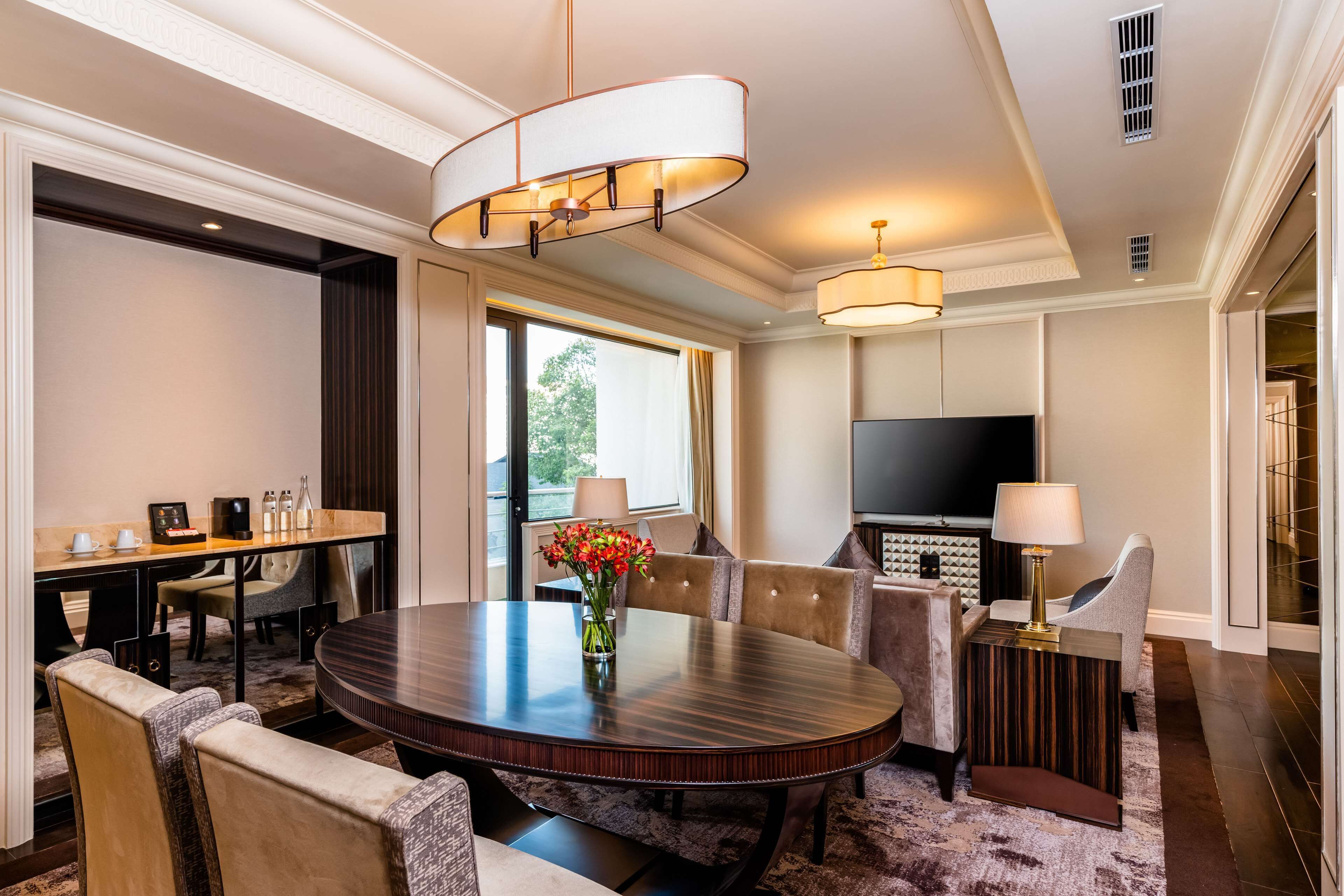 A hotel room with a round dining table, comfortable seating, a flat-screen TV, and a large window.