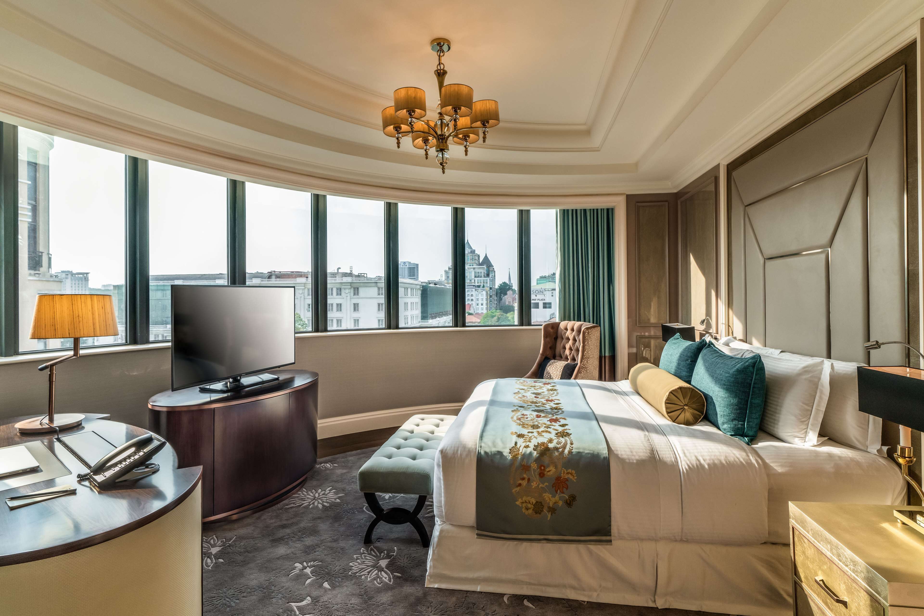 Quarto de hotel com uma cama grande, televisão, escrivaninha com telefone e vista para a paisagem urbana através de amplas janelas.