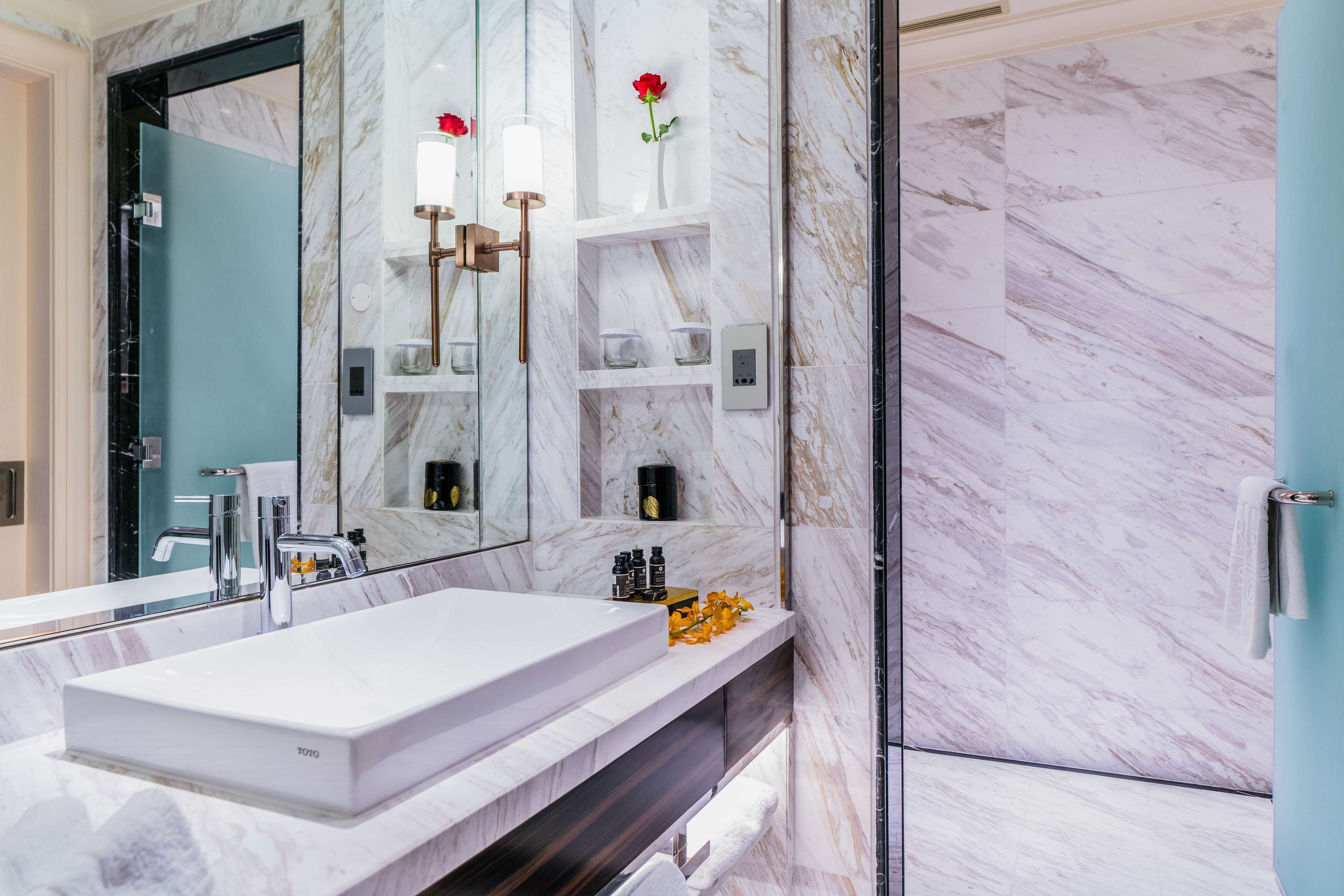 A modern bathroom with a large mirror, a white sink, marble walls, and a shower area.