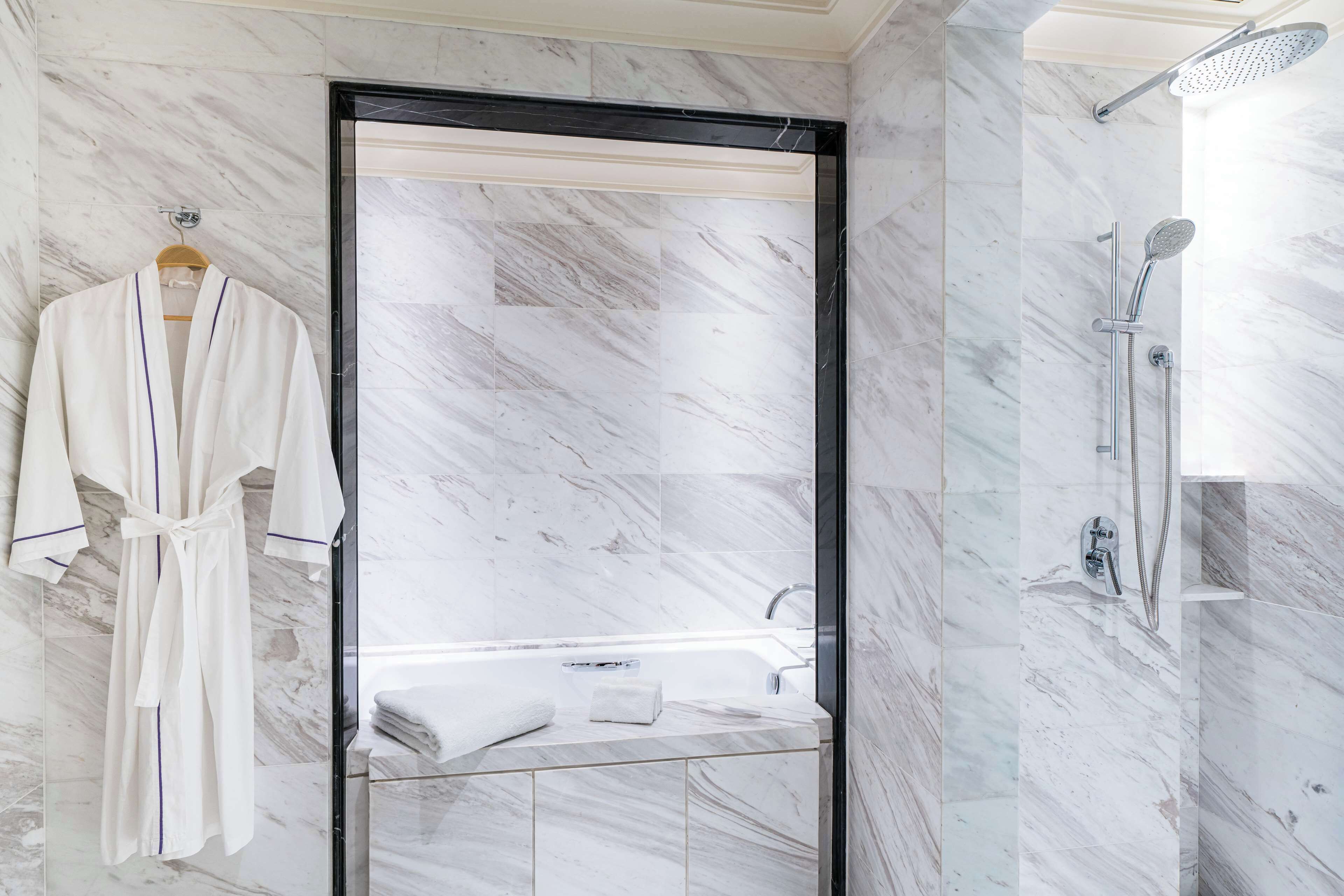Un baño moderno con ducha envuelta en vidrio, una bata blanca colgada de un perchero y pared con azulejos de mármol.