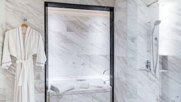 Un baño moderno con ducha envuelta en vidrio, una bata blanca colgada de un perchero y pared con azulejos de mármol.