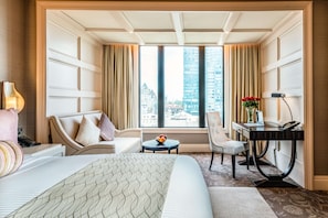 Ein Hotelzimmer mit einem großen Bett, zwei Sesseln, einem Schreibtisch mit Computer, einem kleinen Tisch mit Früchten und Blick auf die Stadt durch große Fenster.