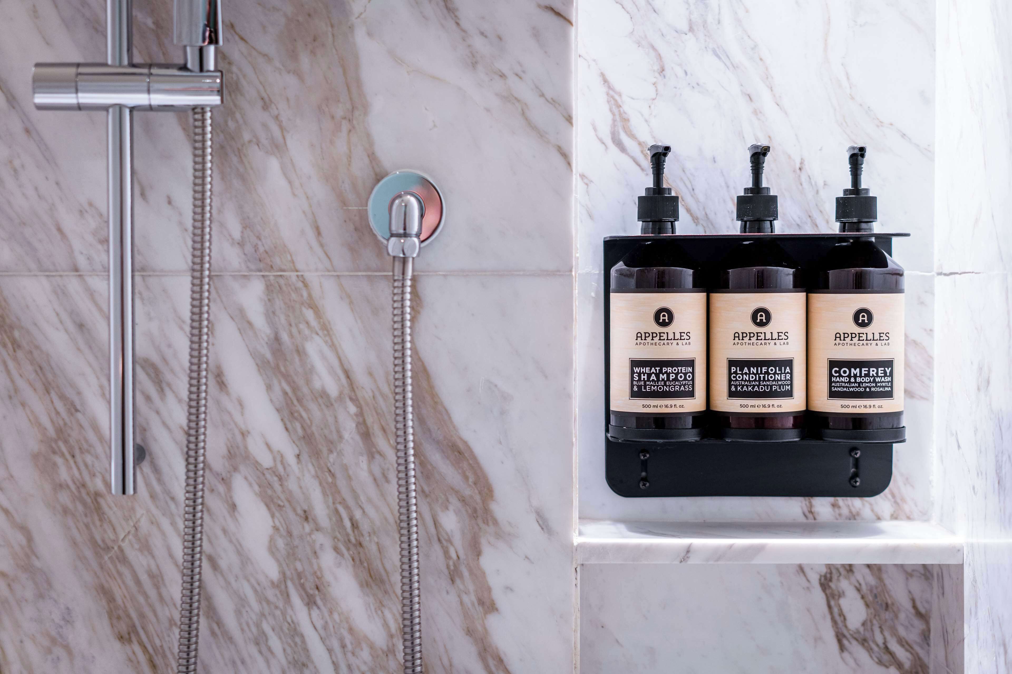 A bathroom with a shower, a marble wall, and a shelf with three bottles of Arpelles products.