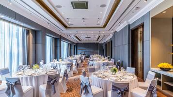 Un salón de banquetes con mesas redondas, sillas y una mesa larga con un escenario al final.